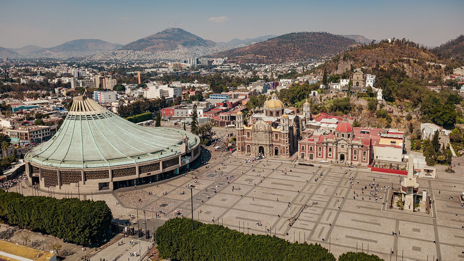 Aerial view of the Guadalupe sanctuary in Mexico City: the round modern basilica with a green roof beside the domed old basilica, a large plaza, and hills behind.