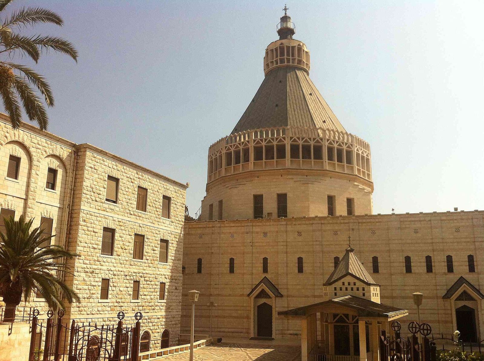 The Basilica of the Annunciation in Nazareth: a pale stone church with a tall cone-shaped dome, beside the convent building, with palm trees in front.
