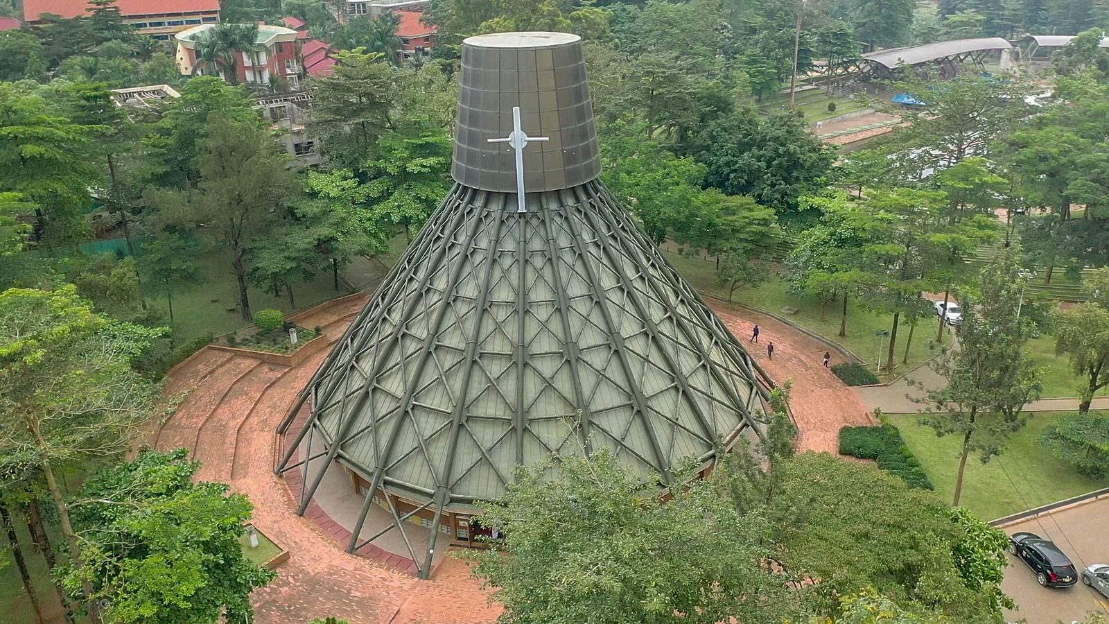Aerial view of the Uganda Martyrs Basilica at Namugongo: a tall cone-shaped roof of steel and glass topped by a cross, among trees.