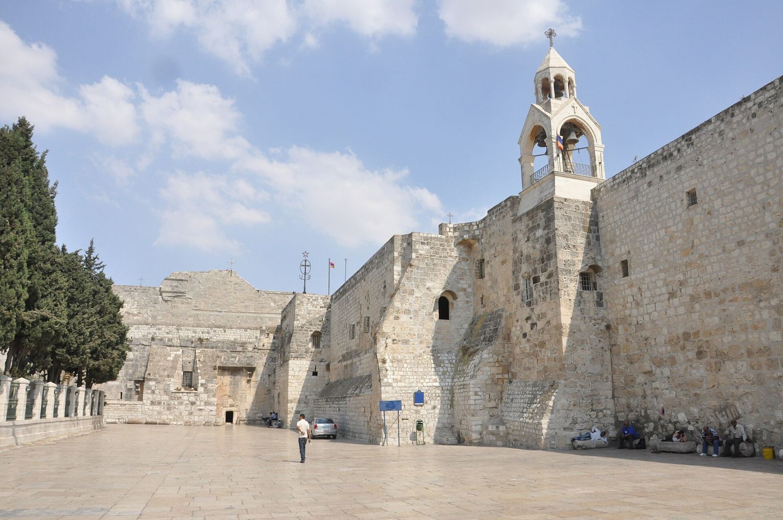 The fortress-like stone front of the Church of the Nativity on Manger Square, with a bell tower, under a blue sky.