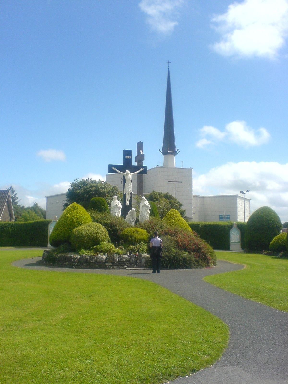 Gardens at Knock Shrine: a shrubbery with a crucifix and white statues on a lawn, a path, and a tall spire and white building behind under a blue sky.