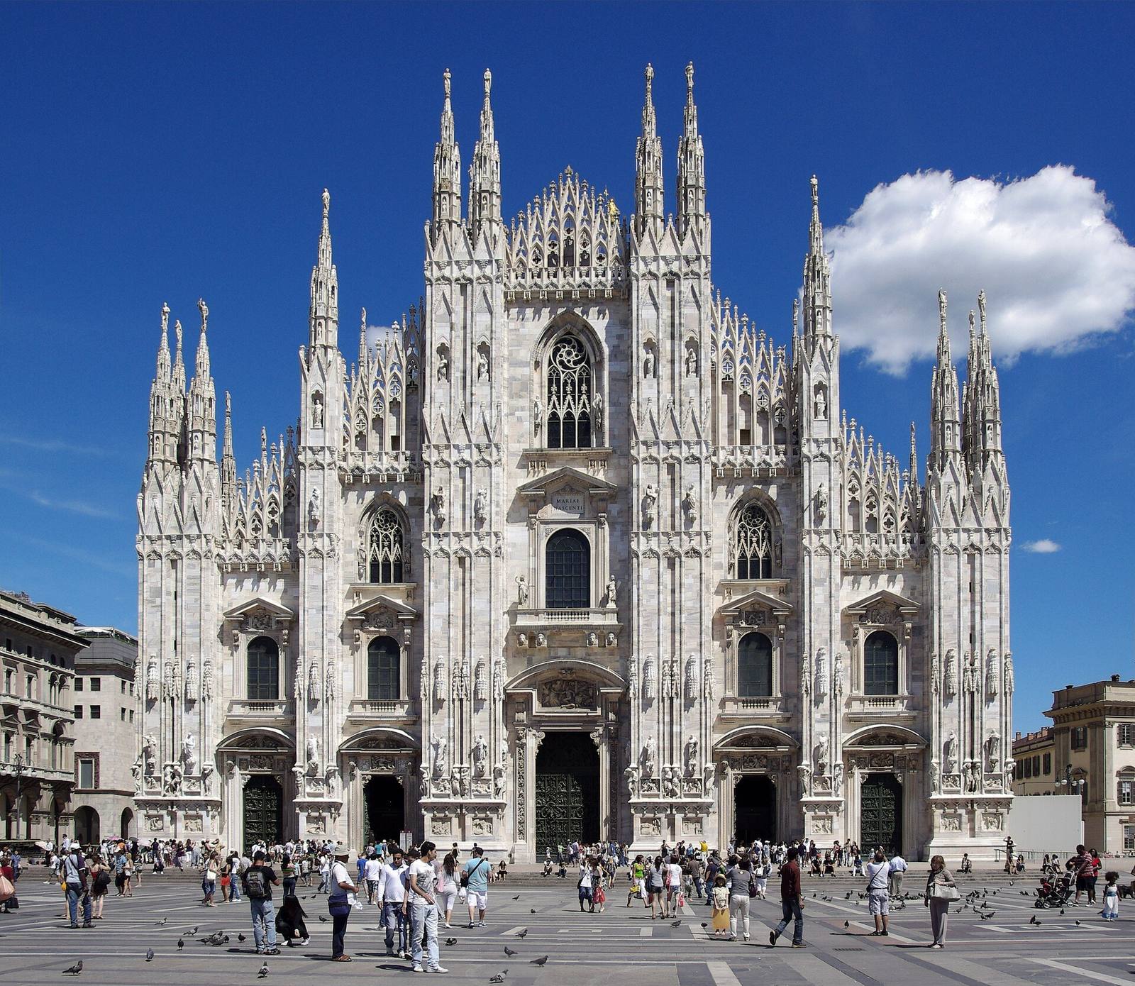 The white marble façade of Milan Cathedral, bristling with spires, above a crowded square.