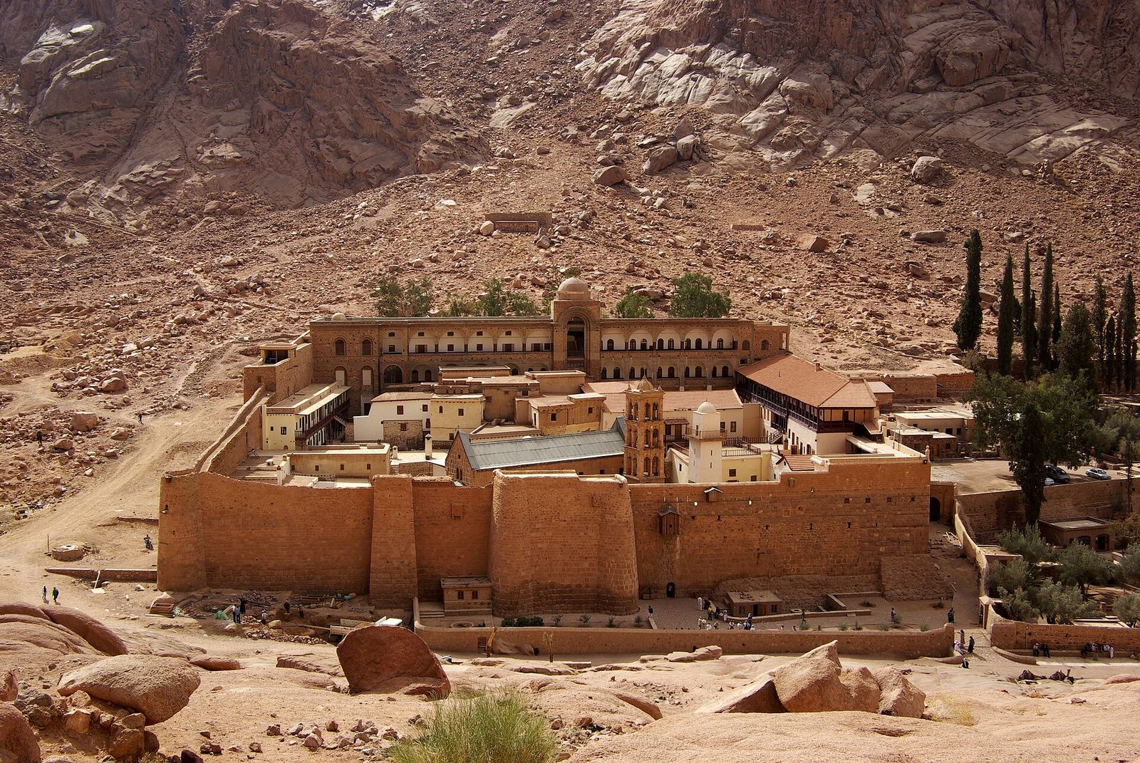 Saint Catherine’s Monastery seen from above: high stone fortress walls enclosing the church and buildings, at the foot of bare red granite mountains.