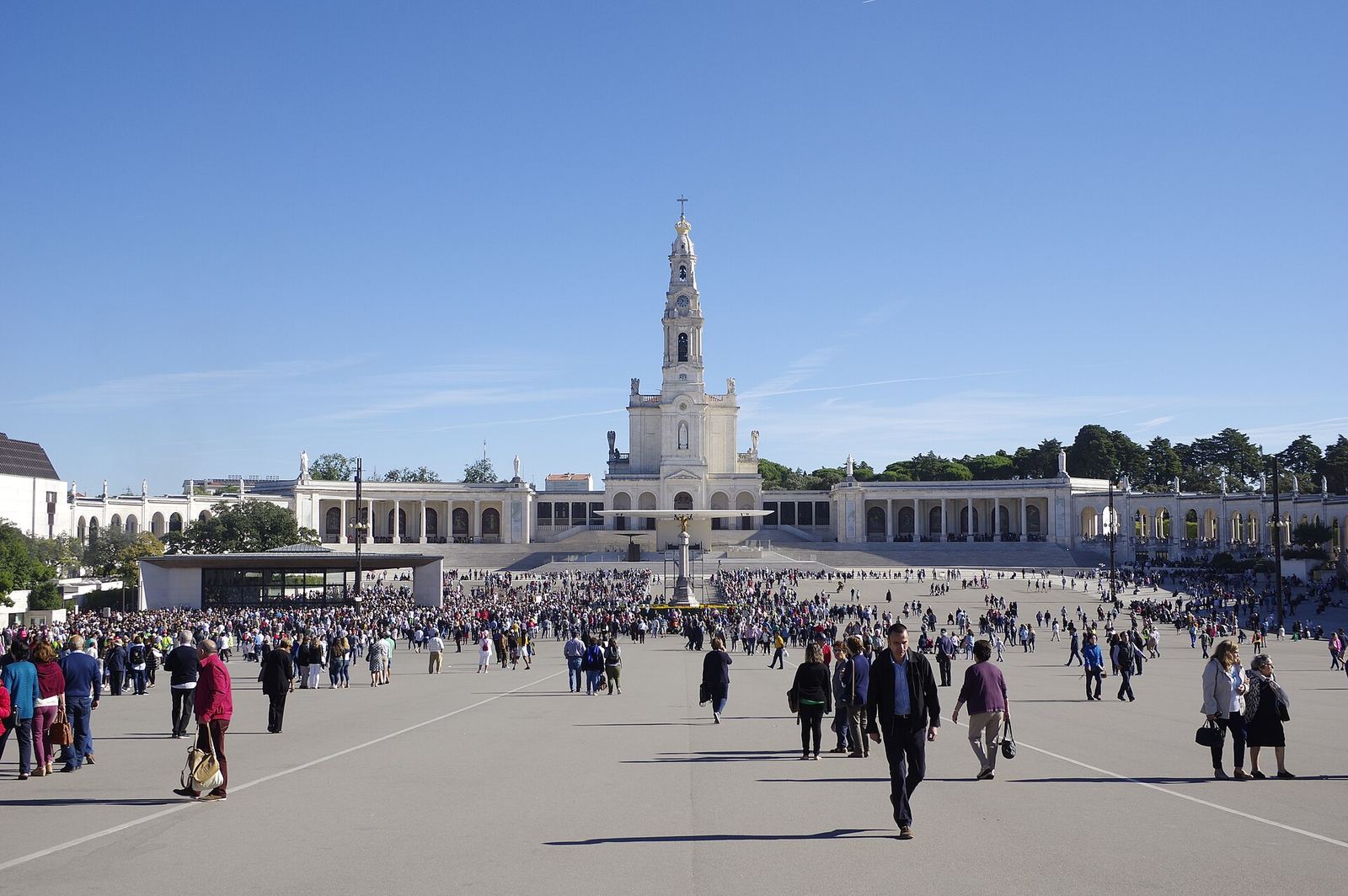 The Basilica of Our Lady of the Rosary at Fátima, white with a tall central tower and curved colonnades, beyond a wide square with pilgrims walking.