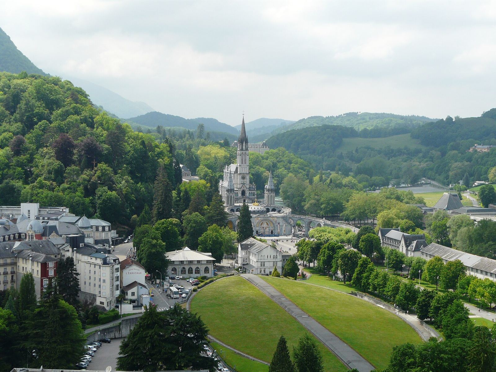 The sanctuary at Lourdes seen from the castle: the spire of the upper basilica rising from wooded hills, with lawns and town buildings in front.